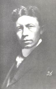  Head and shoulders photograph of a dark-haired man age 30–35, turned towards left but facing camera. His facial expression is serious.