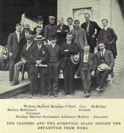 A group of 16 men, standing or sitting, on the deck of a ship with a small lifeboat visible, left background. The group's pose is casual but they are formally dressed, some with hats, others carrying canes.