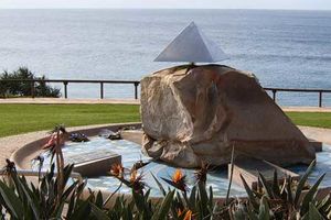 A large stone sitting in a circular basin. The stone is capped with a small metal pyramid. Four plaques also sit in the basin. The entire memorial is located on a hill, with flowers in the foreground and the ocean behind.