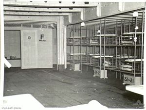 Inside of a medical ward aboard a ship: Bunk beds line the right side, other furnishings protrude into the bottom edge of the photograph, but apart from these, the room is empty. A door at the far end of the room has the text "F WARD" painted on it.
