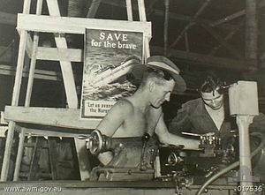 Black and white photograph of two soldiers working with a lathe. A poster behind them depicts a ship with hospital markings sinking by the bow and is captioned with "SAVE for the brave" and "Let us avenge the Nurses".
