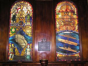 Two tall stained glass windows. The left window shows an aircraft carrier about to launch an aircraft, while the right depicts two cruisers and an aircraft carrier at sea. A memorial plaque sits between the windows.