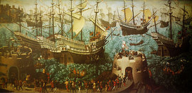 A small fleet of large, highly decorated carracks are riding on a wavy sea. In the foreground are two low, fortified towers bristling with cannon and armed soldiers and retinue walking between them.