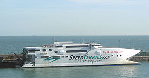 A white catamaran tied up at a wharf. The flank of the ship is decorated with two stylised green arrows near the stern, the word "SPEEDFERRIES.COM" painted along the hull above the words "DOVER-BOULOGNE", and the phrase "Fight the Pirates!" painted in red near the upper bow. Some people are standing on the upper deck, and the open sea can be seen behind the ship