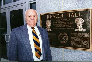 A balding white-haired man stands next to a dedication plaque set into a wall which is entitled "Beach Hall".