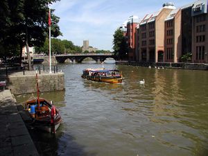 A yellow water taxi on the water between stone quaysides. The far bank has large buildings and in the distance is a three arch bridge.
